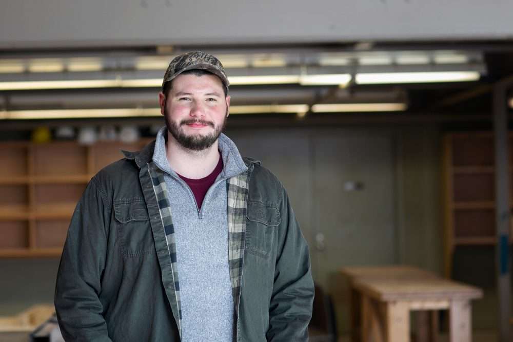 Alumni Spotlight: Charlie Clark stands in a workshop, shelves and wooden table behind him.