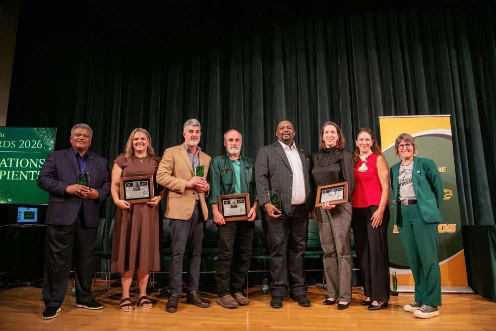 Seven award winners stand on stage with President Bonderoff holding alumni awards, smiling for a group photo.